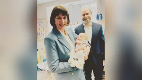 Danielle Cooper Woman on the left wearing a light blue trouser suit with a white blouse and short brown hair is holding a newborn baby who is wearing a cream baby grow and cream boots. Man with a moustache and beard on the right wearing a navy blue suit with a white shirt.