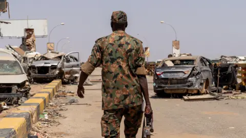 Avaaz via Getty Images A Sudanese soldier holding a gun walks on a street with his back to the camera, with several damaged cars in front of him