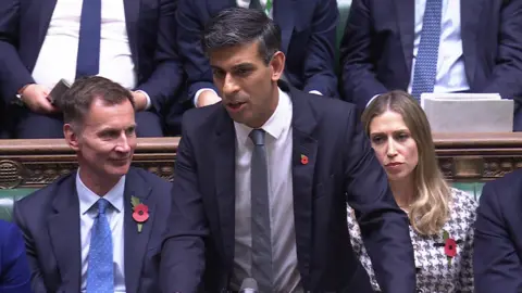 Rishi Sunak wearing a blue suit and a grey tie, standing in the Commons