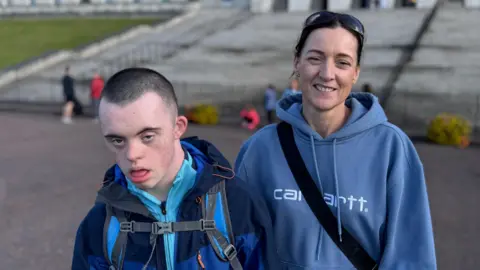 BBC Suzy Ward a její syn Eoyn stojí před velkou krémovou budovou s kamenným schodištěm, které k němu vedlo. Chlapec má hnědé bzučení. Má na sobě světle modrý top s námořnictvem. Má batoh s modrými popruhy. Žena, která stojí k chlapcům, nosí mikinu námořnictva. Má přes hruď popruh černých sáčků.
