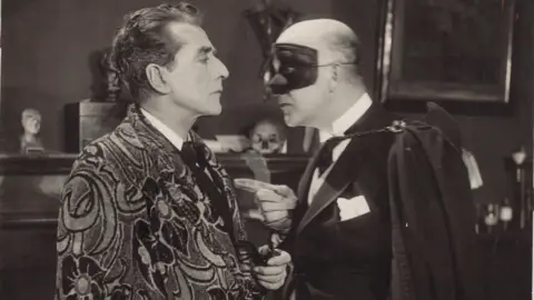 BFI National Archive Black and white photograph of a man in an elaborately patterned dressing gown, confronted by a man wearing a cape, gloves and a opera mask