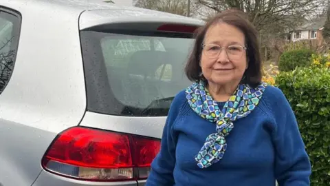 Marbelia Davison pictured outside her home, beside her silver car. Marbelia is wearing a blue jumper, and patterned neckerchief. She has shoulder length hair and is wearing glasses. 