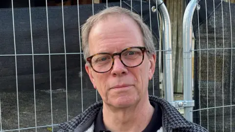 A head shot of Rob Frankson. He is wearing glasses and a black and grey dogtooth patterned coat over a black top. He is outside and silver railings can be seen behind him. 