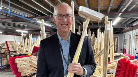 Wiltshire Bobby Van Trust A bespectacled smiling man stands in a large room surrounded by brushes and is holding a brush upside down with his left hand