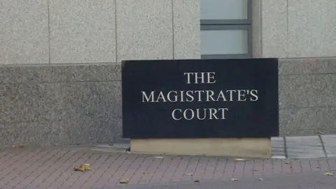 A black, rectangular block with the words 'The Magistrate's Court' in capital letters. It sits on a concrete plinth and is displayed in front of a grey building. Paving stones are visible beneath it.