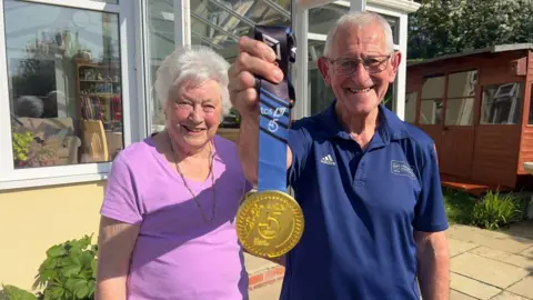 John Fairhall/BBC Ros (left) in purple top smiles as Mac (right) in blue polo shirt holds up his medal. They are standing in a garden and both are smiling.