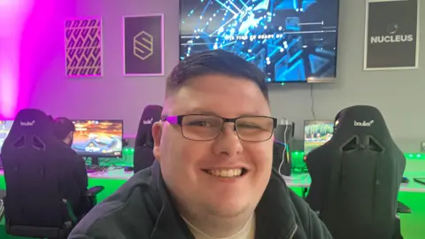 Kyle Faulkner smiles, he has black glasses. Behind him there are gaming chairs and gaming monitors. There is a large monitor on the wall.