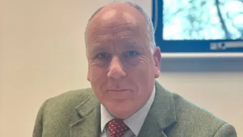 Paul Clifton looking at the camera, wearing a green jacket, red tie and blue and white shirt.