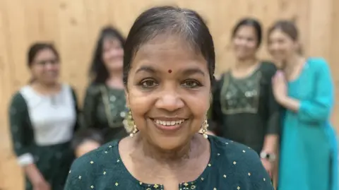 Head and shoulders photograph of Anne Tiburtius. Anne is wearing a green tunic with gold detail, she is smiling with a bindi on her forehead.
