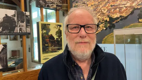John Devine/BBC Richard Carter has wispy light brown hair with matching beard and moustache, he has a black fleece on with dark checked opened neck shirt underneath, he has glasses on with a black frame, behind him are displays in a museum.