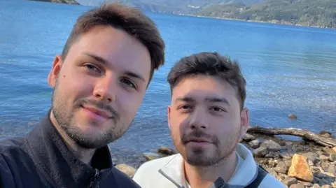 Joe Diaz and Dan Campbell. Both have brown hair. Joe is wearing a black jumper and Dan a white jumper. They are standing in front of a large body of water. 