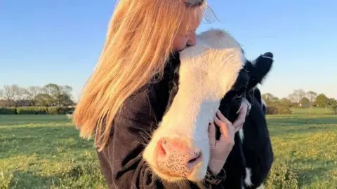 Alice Grammer A woman with long blonde hair hugging a black and white cow. They are in a grassy field. Se is wearing a black fleece.