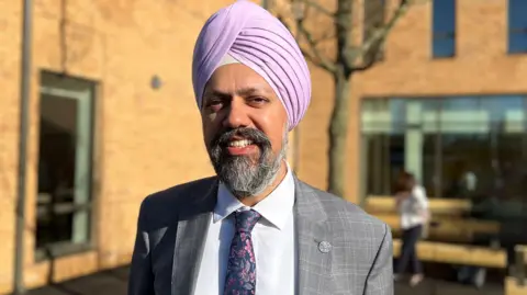 Tan Dhesi is smiling and wearing a grey suit with a white shirt. He has a tie with a navy background and pink flowers. He is also wearing a light pink turban. In the background the new building is visible. 