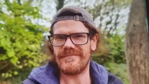 Devon and Cornwall Police Shane Lynch pictured wearing a navy blue hoodie and navy blue jacket. He is also wearing a grey cap and has black square glasses. He also has long ginger hair and a ginger beard.