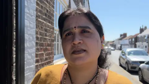 A woman with dark hair, tied back, is wearing a yellow sari and a beaded necklace as she stands in a street outside a brick building. Cars are parked to the right and houses can be seen in the background.