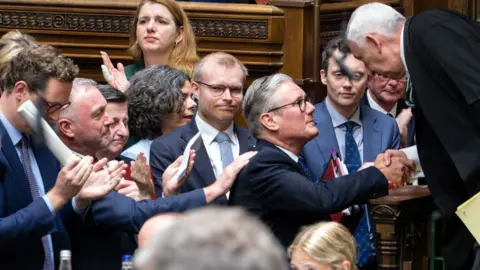 Keir Starmer shakes hands with Lindsay Hoyle, Speaker of the House of Commons, as people clap and pat Stamer on the back after his last PMQ session as Prime Minister