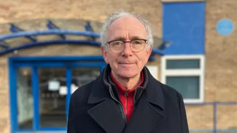 Vernon Orr in front of The King's Centre on Osney Mead. He is wearing glasses and has a slight smile. 