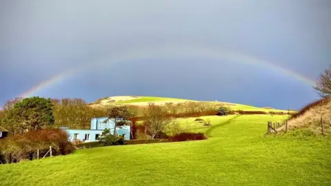 A rainbow spans the dark grey sky mirroring the shape of a green hill beneath it with a blue house nestled into the lea of the slope surrounded by trees