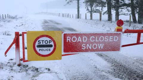 Peter Jolly/Northpix The metal gates are closed across a snow-covered road. There are signs on the gate that warn: "Stop. Police" and "Road closed. Snow".