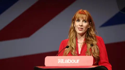 Angela Rayner addresses the Labour Women's Conference in Liverpool last year. She stands at a podium with Labour's red rose logo, wearing a bright red dress, in front of a Union Jack flag. She wears her long hair loose and styled into curls.