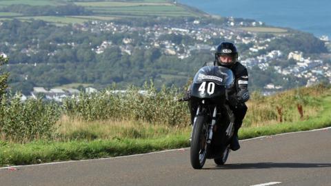 Ian Bainbridge, wearing black leathers, rides his Manx Norton, numbered 40, out of Ramsey on the TT Course