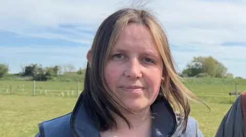 A woman with brown, shoulder-length hair and brown eyes looks at the camera. She is wearing a blue jacket and standing in a green field under a blue, cloudy sky.