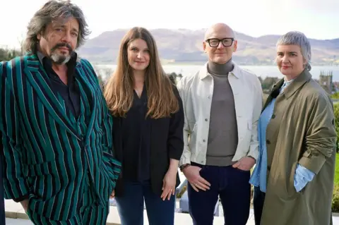 Laurence Llewelyn Bowen in a stripey green and black suit with three unknown others - a woman in dark clothing and long brown hair, a bald man in a white denim jacket and blue jeans and a grey shirt, and a woman with a short haircut wearing a green trench coat and denim blue shirt, pictured outside with mountains in the background