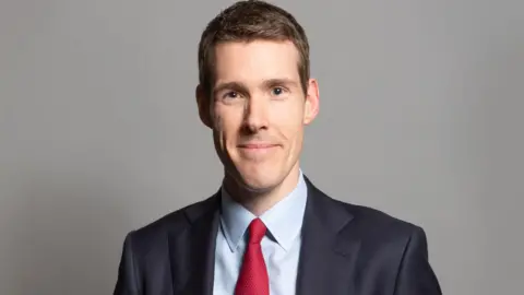 UK Parliament A professional head and shoulders photo of a man with short blonde hair and wearing a black suit with a red tie. He's standing against a grey background and smiling at the camera.
