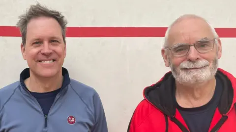 Annabel Amos/ BBC A man in a blue zip top stands next to a man with glasses and a beard in a red zip top both smiling at the camera. 