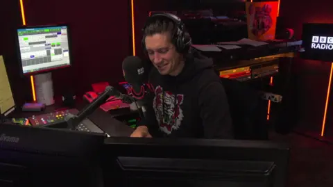 Vernon Kay sits in a studio next to a microphone. He is wearing a black hoodie, and smiles as he interviews Arthur about the missing gnomes. 