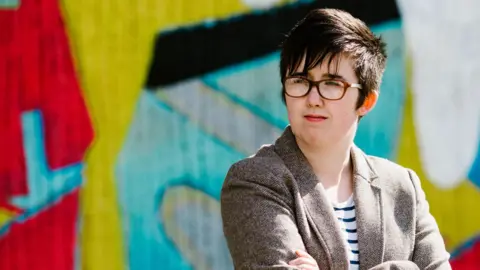 Lyra McKee stands with her arms folded in front of a brightly painted wall. She is a young woman with short black hair, with a fringe over her forehead coming to rest on her glasses. She is smiling She is wearing a brown tweed jacket over a white a navy striped top.