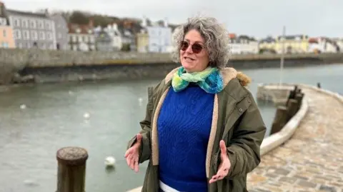 A woman in sunglasses with a blue guernsey jumper, green coat and green and blue scarf on, speaks to a person off-camera while standing on a marina with the ocean and seafront in the background.