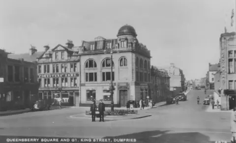 Heritage Service, Dumfries and Galloway Council A black and white picture of Queensberry Square and Great King Street in the centre of Dumfries. Wide open roads with just a handful of cars. Two policemen are standing in the middle of the square. 