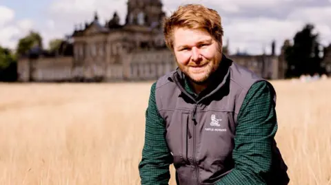 Emma Raye A man, Guy Thallon, wears a green and navy chequered shirt and a Castle Howard branded gilet. He is kneeling in a field with the large historic house in the background.