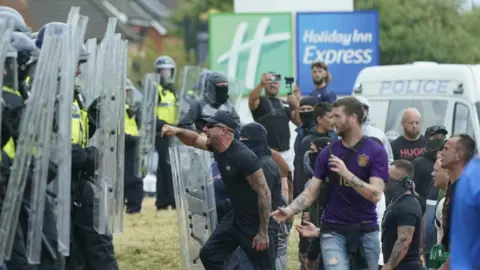 PA Media Angry young men shout in the face of a row of police officers in riot gear