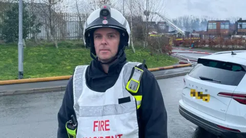 A man called Martin Sleigh stands in front of the scene of a school fire in Devon. He is a firefighter and is wearing his firefighting gear. He has a white high-vis overall on. He also has a white helmet on.