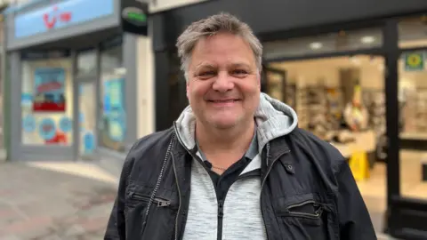 Paul Pinel is smiling at the camera. He is in a dark jacket, with a grey hoodie underneath and is standing outside in front of some shops.