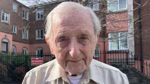 BBC Shows an elderly man in a light shirt and white t-shirt standing in front of a building