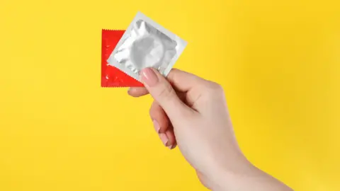 A woman's hand holding up a red and a silver condom packet, in front of a yellow background.