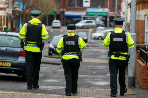 Three police officers in yellow jackets and dark vests walking down a street with parked cars in front of them