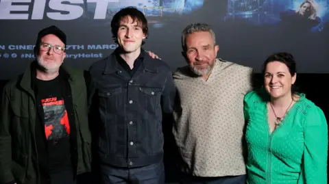 Darren Kidd PressEye Four people stand in a row. The man on the left has glasses and is wearing a red and black T-shirt with a black jacket and black hat. The man second from the left is taller and wearing a black shirt and has dark hair. The man second from the right has grey/blonde hair and beard and is wearing a patterned beige top. A woman stands at the far right. She has dark hair and is wearing a green top.