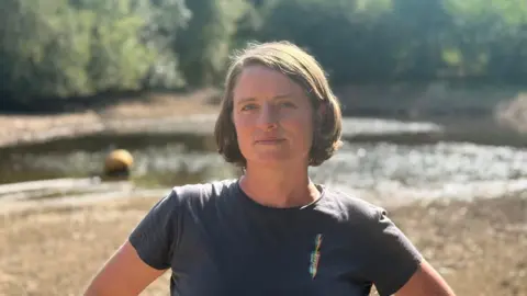 Riverford Harriett Bell stands in a dry reservoir at Riverford Organic Farmers. She has a brown bob and is wearing a blue T-shirt. There is small amount of water in the reservoir, with the sun glinting off it. 
