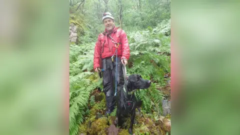 Keswick Mountain Rescue Team Man in red jacet and white helmet with a black dog in his lead - both are soaked from the rain