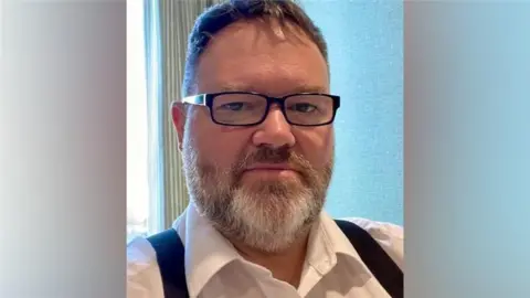 Anton Goodwin A man with short brown hair, greying beard and black-framed glasses is wearing a white shirt and black braces. He is sitting in a room with a pale blue wall and light-striped curtain by a window.