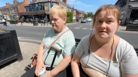 Two women sit on a bench. Charlott'e has brown hair and wears a white top. Her mum has blonde hair, a walking stick and is wearing a pale green top with a tan handbag over her left shoulder.