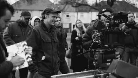 Hanna-Katrina Jędrosz A black and white photo of a man wearing a denim jacket, baseball hat and headphones. Beside him is a woman in a beanie operating a large camera. Behind them, about half a dozen other people look on. There are also houses in the background.