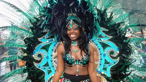 A woman wearing an elaborate carnival costume with blue gemstones, green and black feathers, and a feathered headdress.