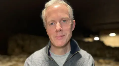 A man is smiling, he has short grey hair and is wearing a grey and black, zip-up jumper. He is standing in a dark room. 