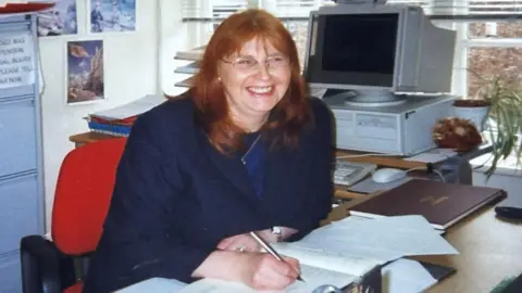 Anne Robson Old photo of Anne smiling while sitting at her desk signing paper work with a silver fountain pen. She has shoulder length red hair and is wearing glasses and a navy suit. Behind her is a very old style computer.