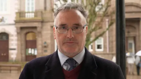 A person wearing glasses, a dark coat and tie stands outdoors in front of a historic stone building. The background is softly blurred, with a tree and a few people holding umbrellas visible on a rainy day.